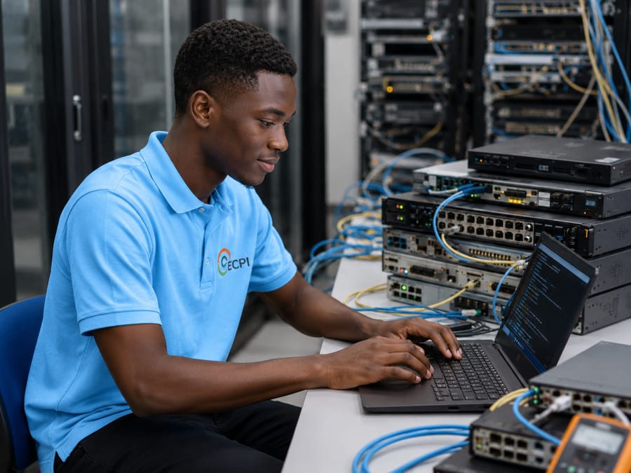 Étudiant en polo bleu configurant un commutateur depuis son ordinateur portable, dans la salle réseau de l'ECPI Dakar